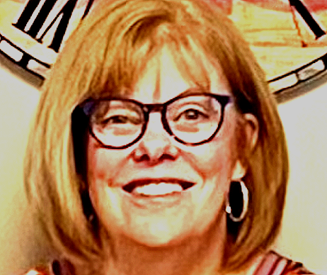 A woman with red hair wearing black glasses and hoop earrings, smiling in front of a clock.