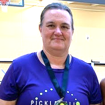 A woman wearing a blue T-shirt with the word 'PICKLE' on it, standing indoors with a yellow wall in the background.