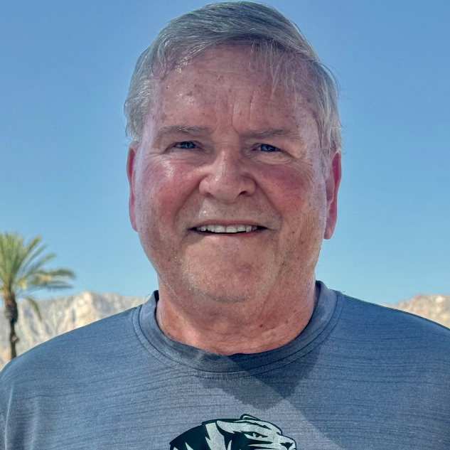 A middle-aged man with short gray hair smiling outdoors against a clear blue sky, with palm trees and mountains in the background, wearing a blue t-shirt.