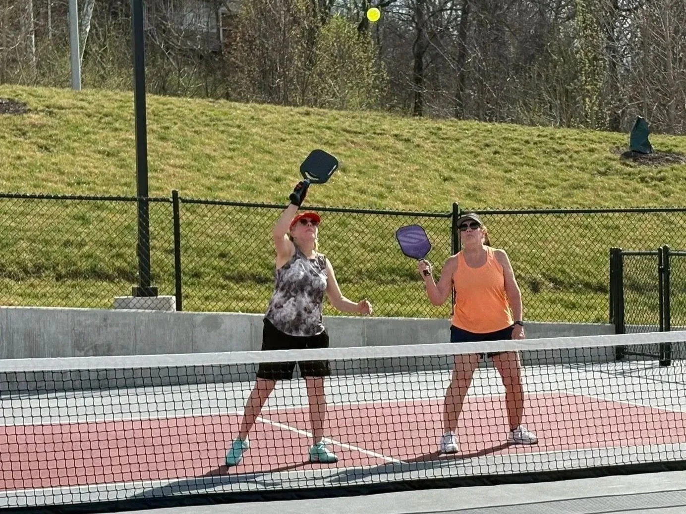 Two women are playing pickleball on an outdoor court, one woman is about to hit a yellow ball with a paddle and the other holds a paddle in her hand, both are wearing sunglasses and sporty clothing.
