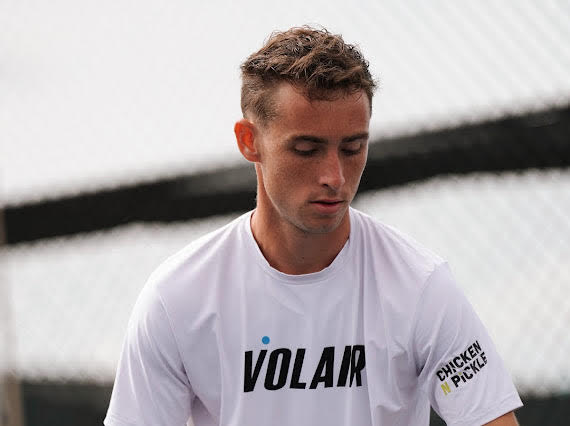 A young man with short, curly hair looking downward wearing a white sports t-shirt with 'VOLAR' written on it and a patch that says 'CHICKEN N PICKLE' on the sleeve.