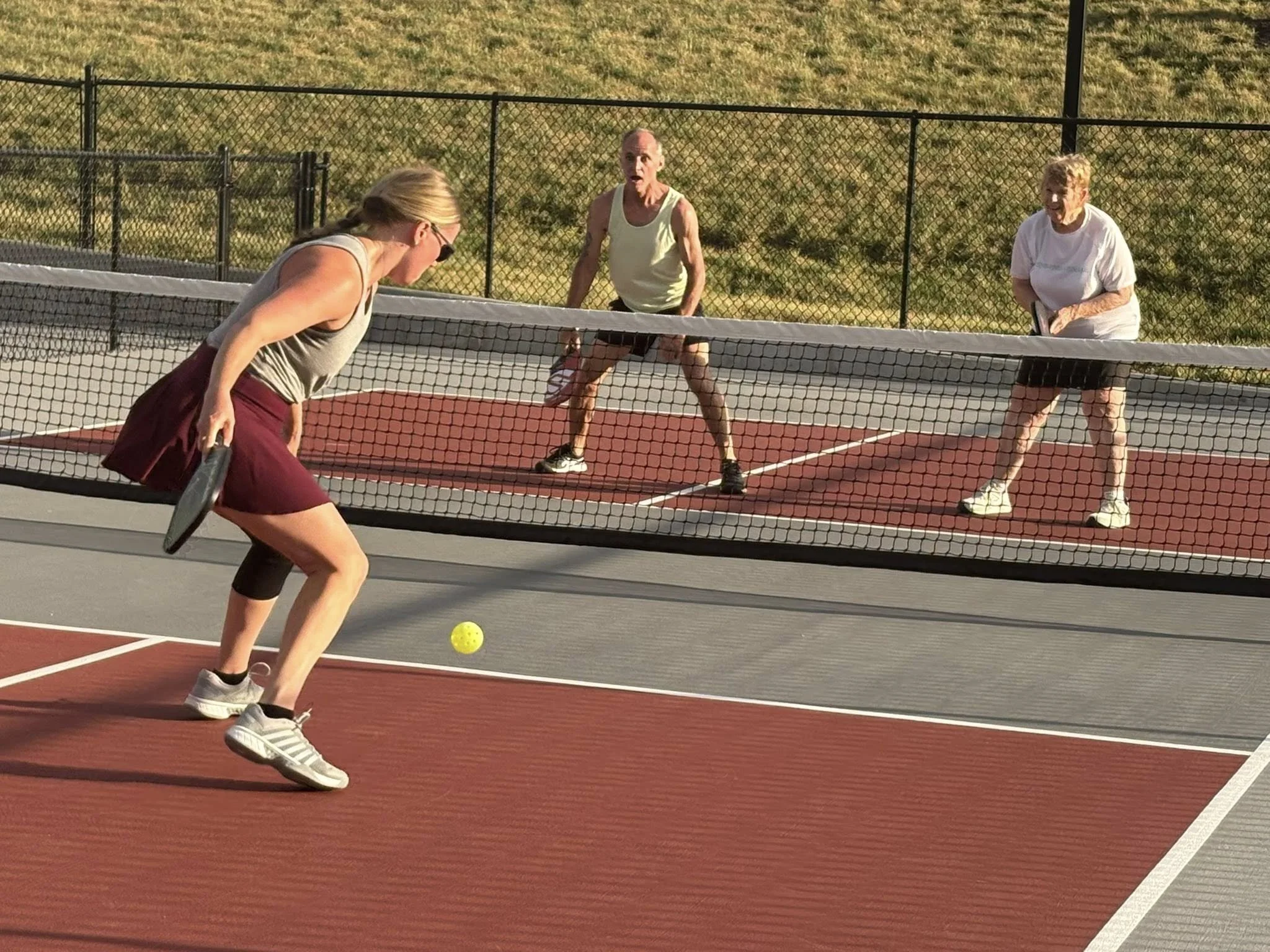 Two women are playing pickleball on an outdoor court, with a net between them and a grassy field in the background. One woman is in the foreground, preparing to hit a yellow pickleball, while the other woman is on the opposite side of the net, ready to receive.