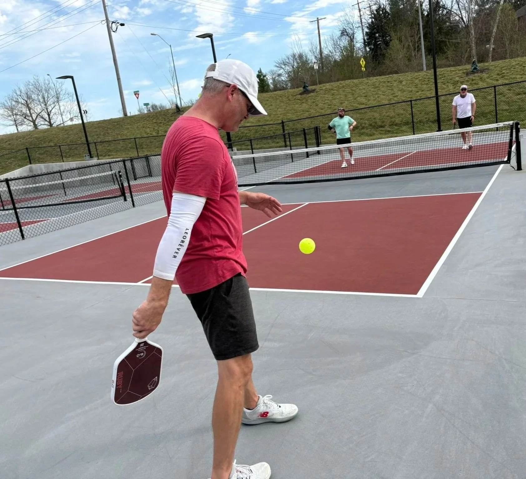 A man in a red shirt, black shorts, white cap, and white sneakers stands on a tennis court holding a tennis paddle, with a yellow tennis ball nearby. In the background, three other men are practicing or playing tennis on the same court, which is outdoors with a green grassy hill, trees, and utility poles behind a black fence.