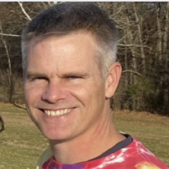 A smiling man with short gray hair outdoors in a grassy area with trees in the background.