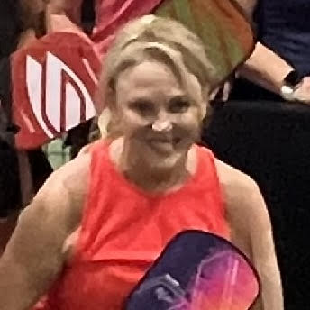 A smiling woman in a red tank top holding a purple and pink tennis racket at a tennis match.