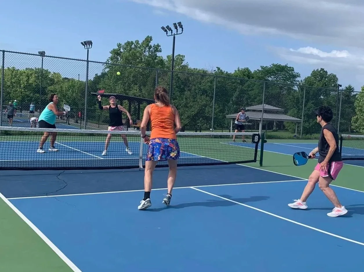 People playing pickleball on an outdoor court, with four players actively engaged in the game and a background of trees and a pavilion.