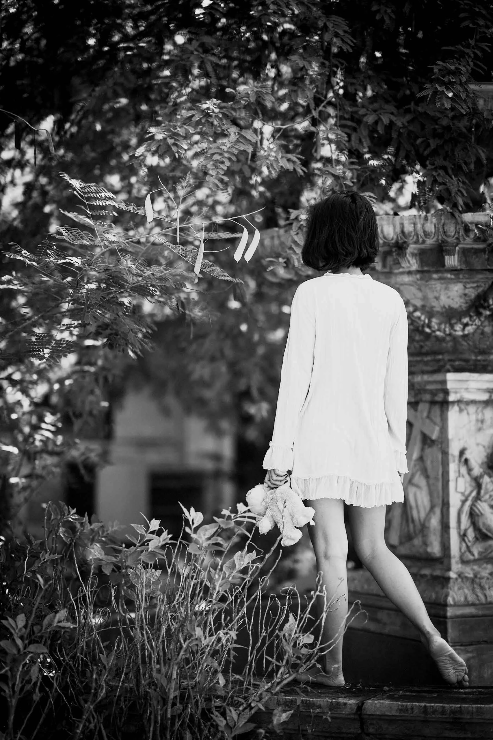 A black and white photograph of a woman in a long, frilly dress standing barefoot on a wooden deck, surrounded by foliage and trees, with her back facing the camera.