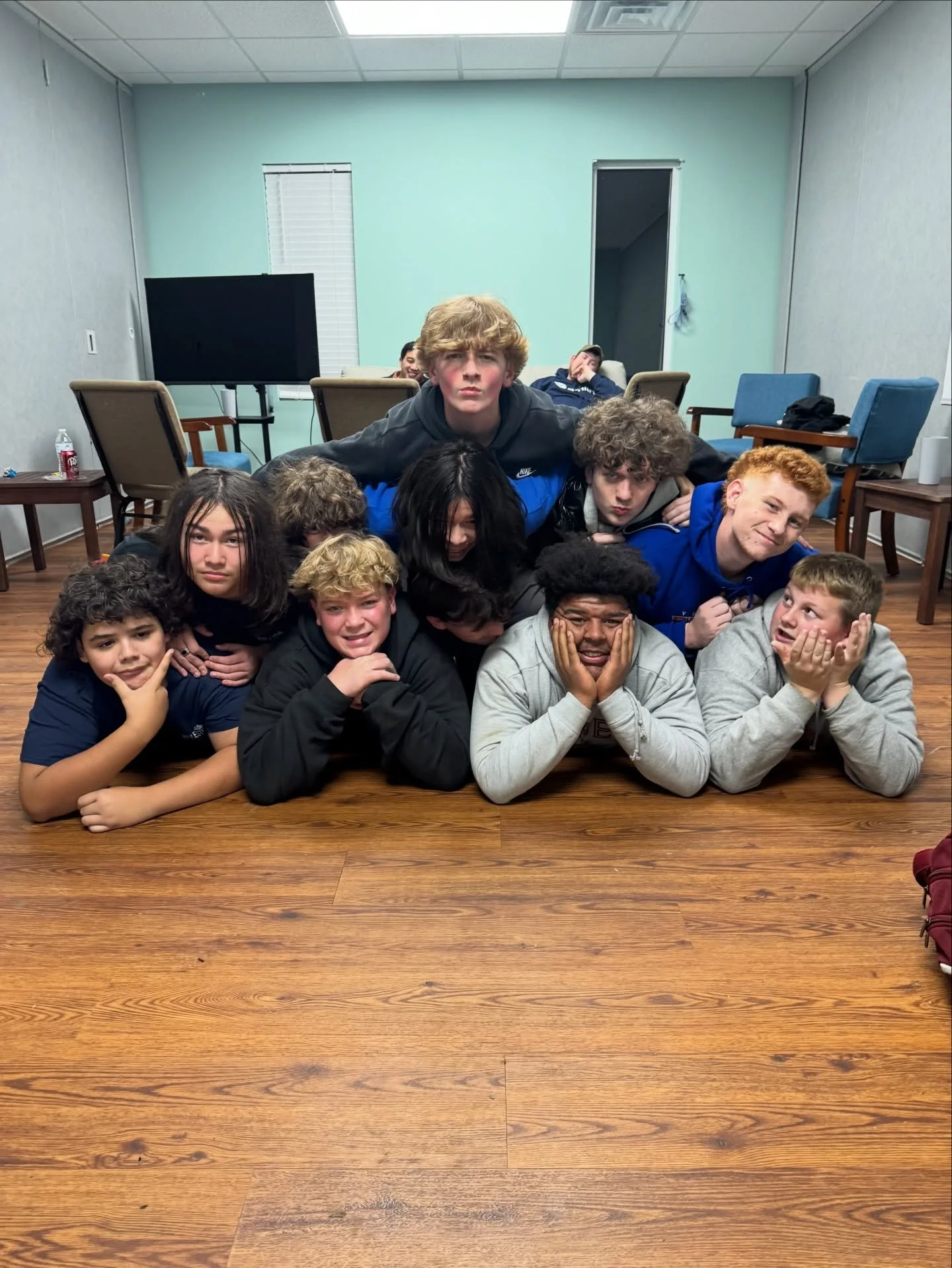 Group of young people gathered closely on a wooden floor in a room with chairs and a television in the background, making funny faces and poses.
