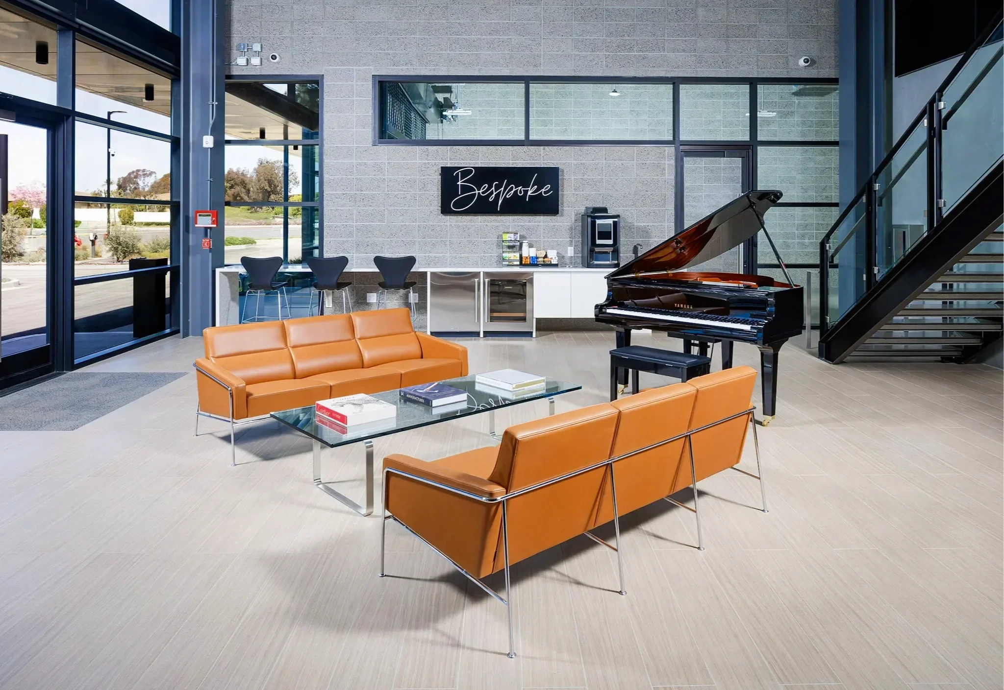 Modern lounge area with orange leather sofas, glass coffee table, grand piano, black chairs, and a sign that reads 'Bespoke' on the wall, large windows, and staircase.