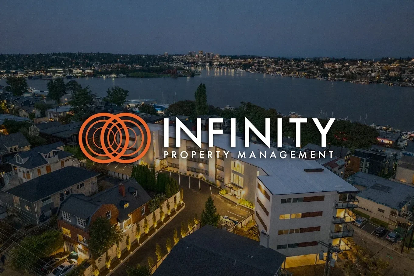 An aerial view of a modern apartment complex at dusk with a river and city skyline in the background, overlaid with the logo and text for INFINITY PROPERTY MANAGEMENT.