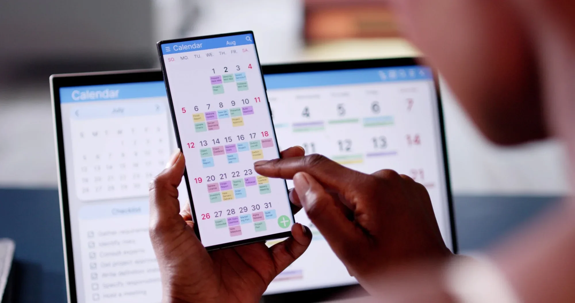 Person holding a smartphone showing a colorful calendar app, with additional calendars on laptop screens in the background.