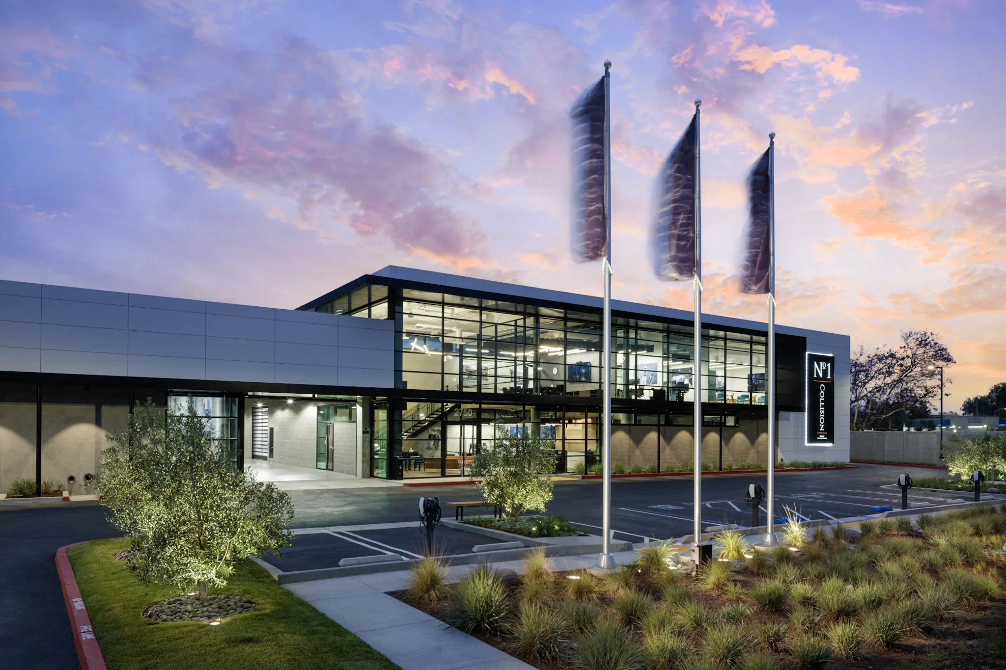 Modern building with glass exterior and three flagpoles with dark flags at sunset, with trees and landscaping in the foreground.