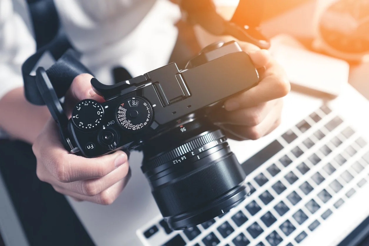 Close-up of a person holding a camera over a laptop keyboard, with some papers and glasses in the background.