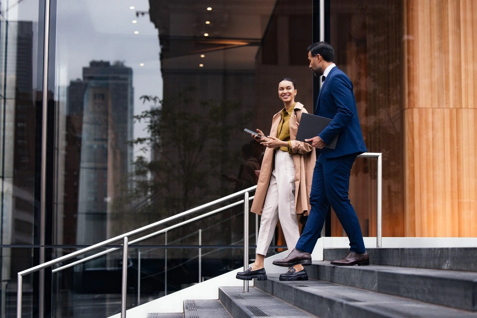 Two business professionals, a man and a woman, are walking down the steps of a modern office building. The woman is smiling and looking at her phone, while the man is holding a laptop and appears to be talking to her. The building has large glass windows and a wooden exterior.