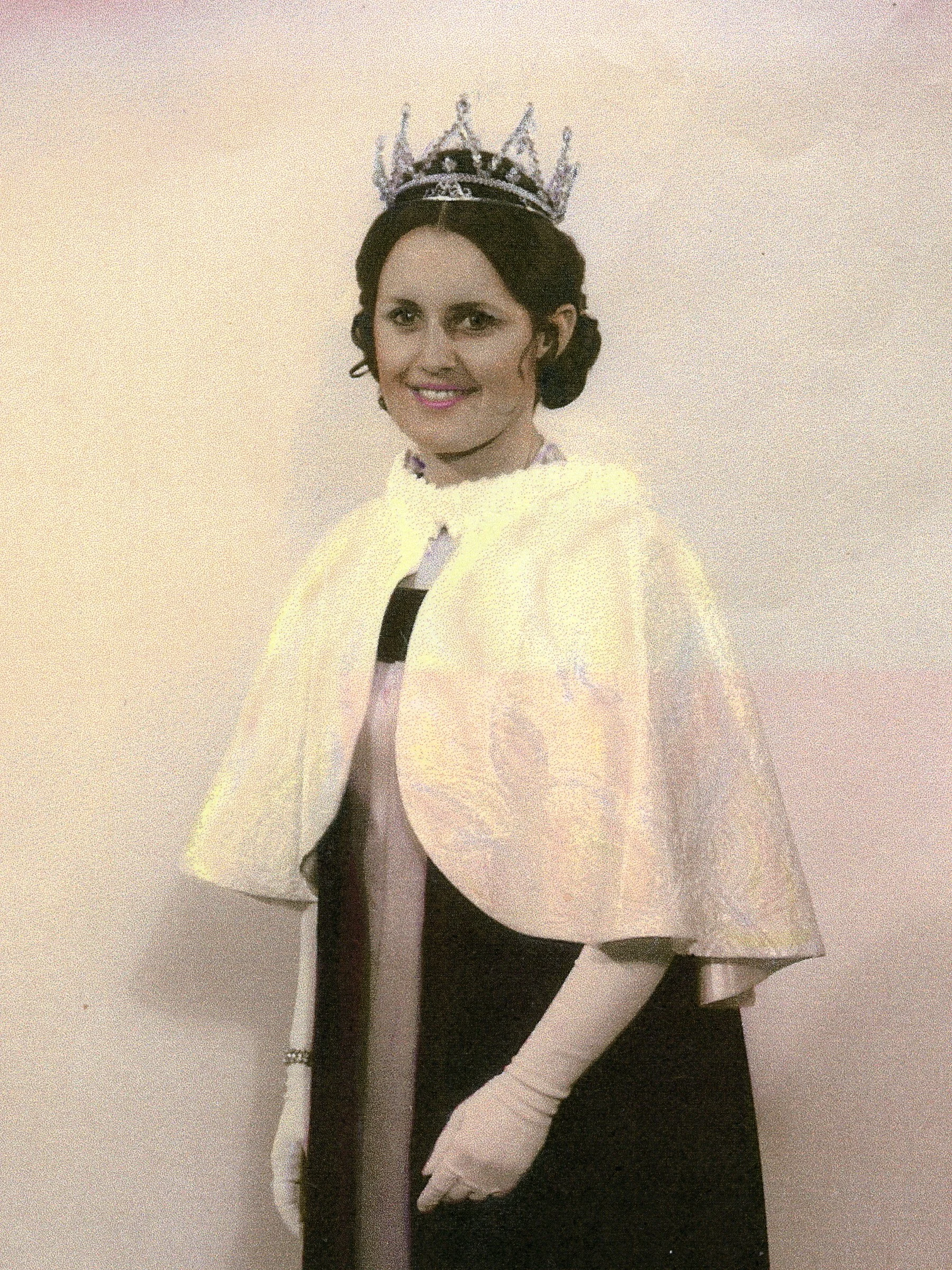 A woman wearing a princess crown and a formal black and white gown, standing against a plain wall.