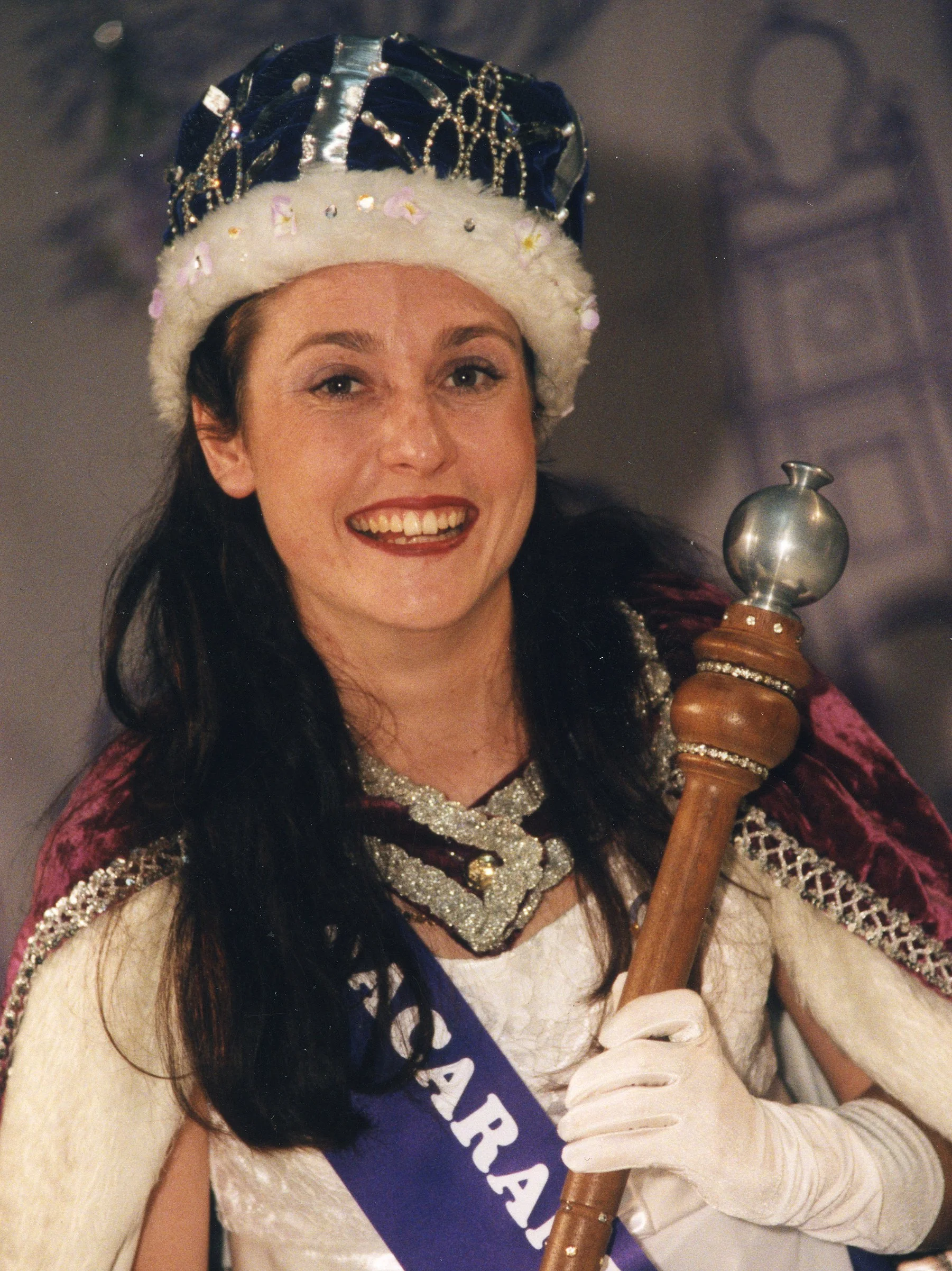 A woman dressed as a queen with a crown, a sash reading 'Queen,' and holding a scepter, smiling.