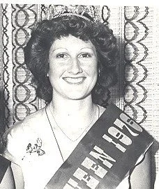 A smiling woman with curly hair holding a sign that says 'U.S. HOUSE' in front of a patterned curtain.