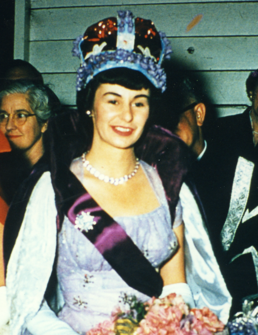 Woman wearing a crown and sash, smiling at a celebration, with other people in formal attire around her.