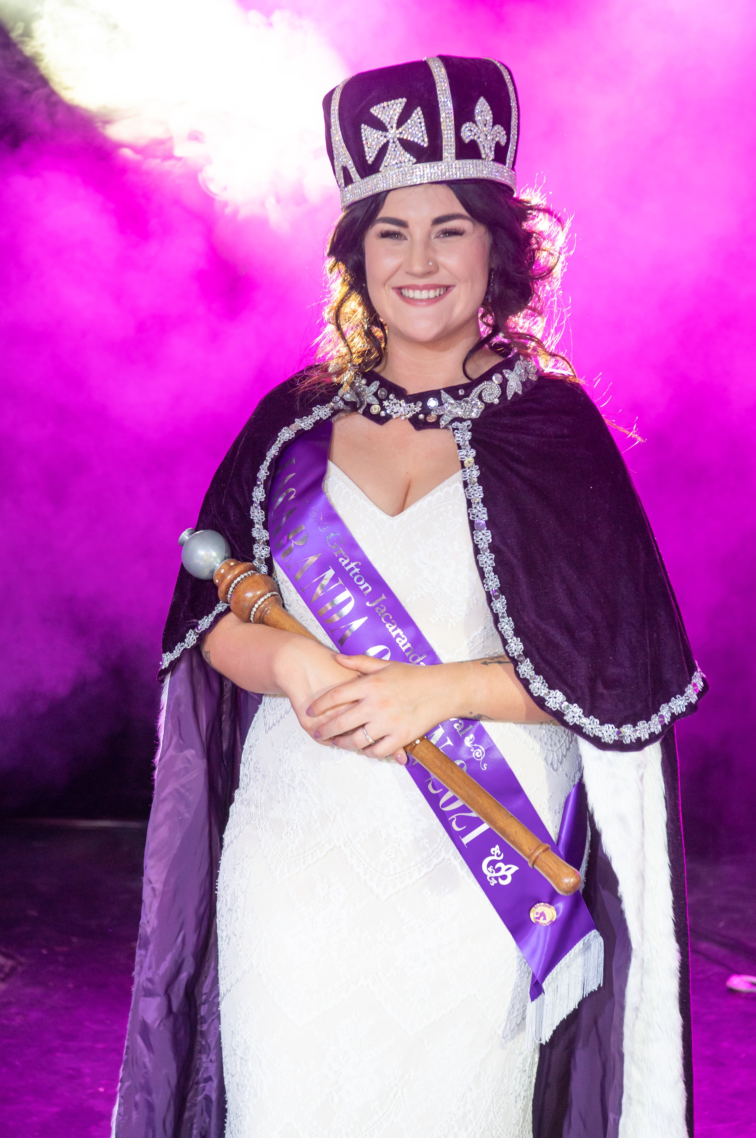 A woman in a white dress and black cloak with silver trim, wearing a purple sash, holding a wooden scepter, and wearing a large jeweled crown, standing against a pink and purple smoky background.