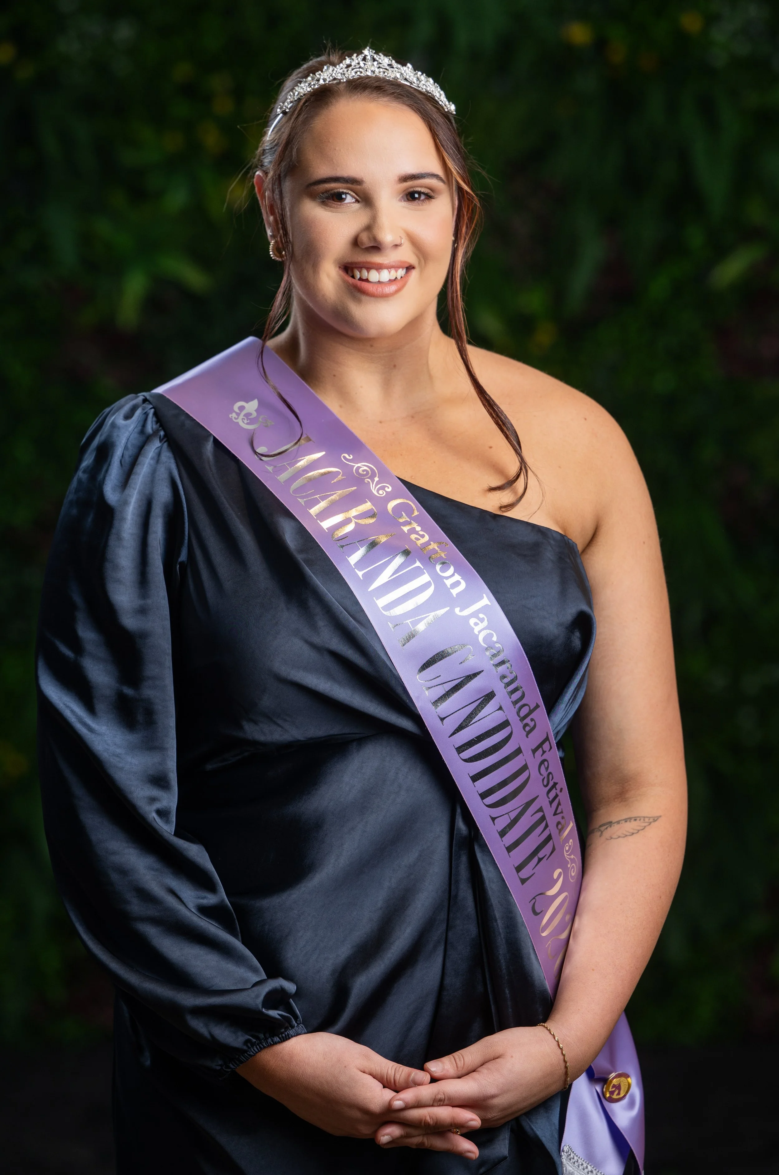 A young woman wearing a tiara, black satin gown, and purple sash with gold lettering, standing outdoors with a blurred green background, smiling at the camera.