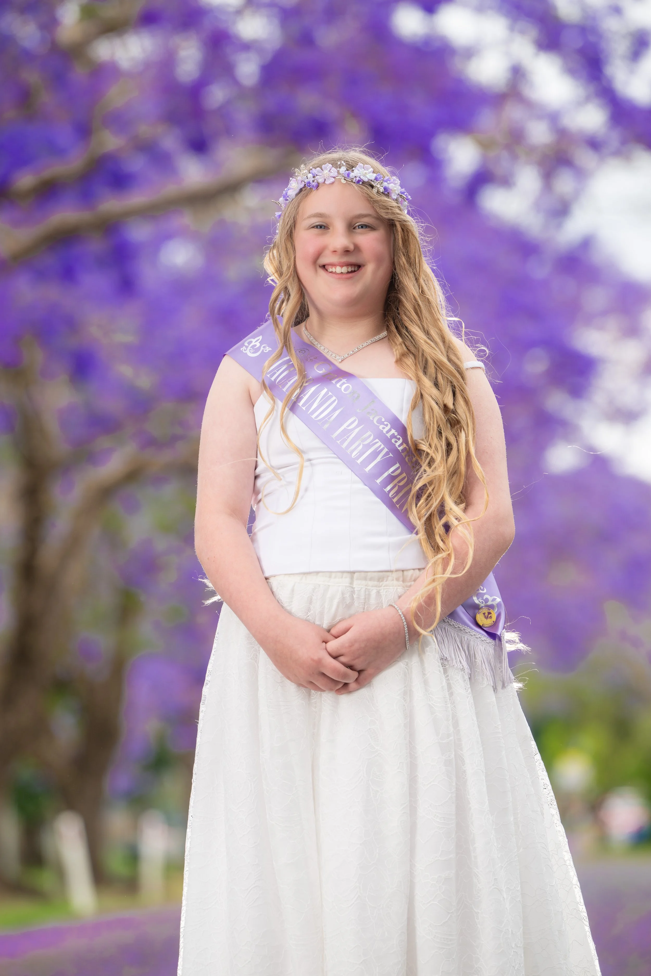 A young girl with long, wavy blonde hair wearing a white dress, a floral crown, and a purple sash that says 'Maldana Valley Princess,' standing outdoors with a background of purple flowering trees.