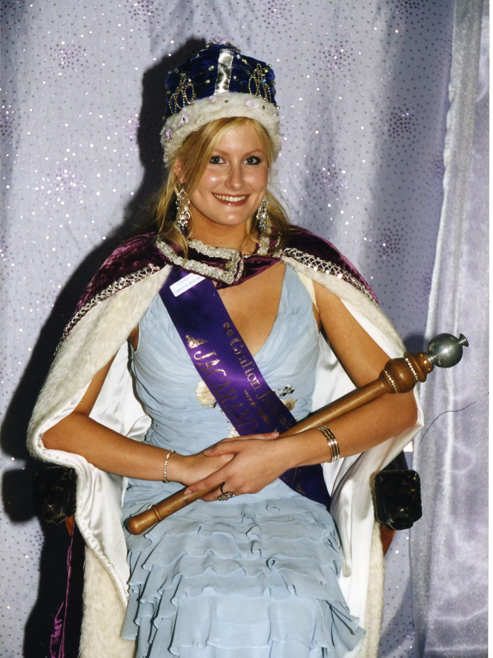 A woman dressed as a queen or princess, sitting on a throne, wearing a crown, sash, and a cape.