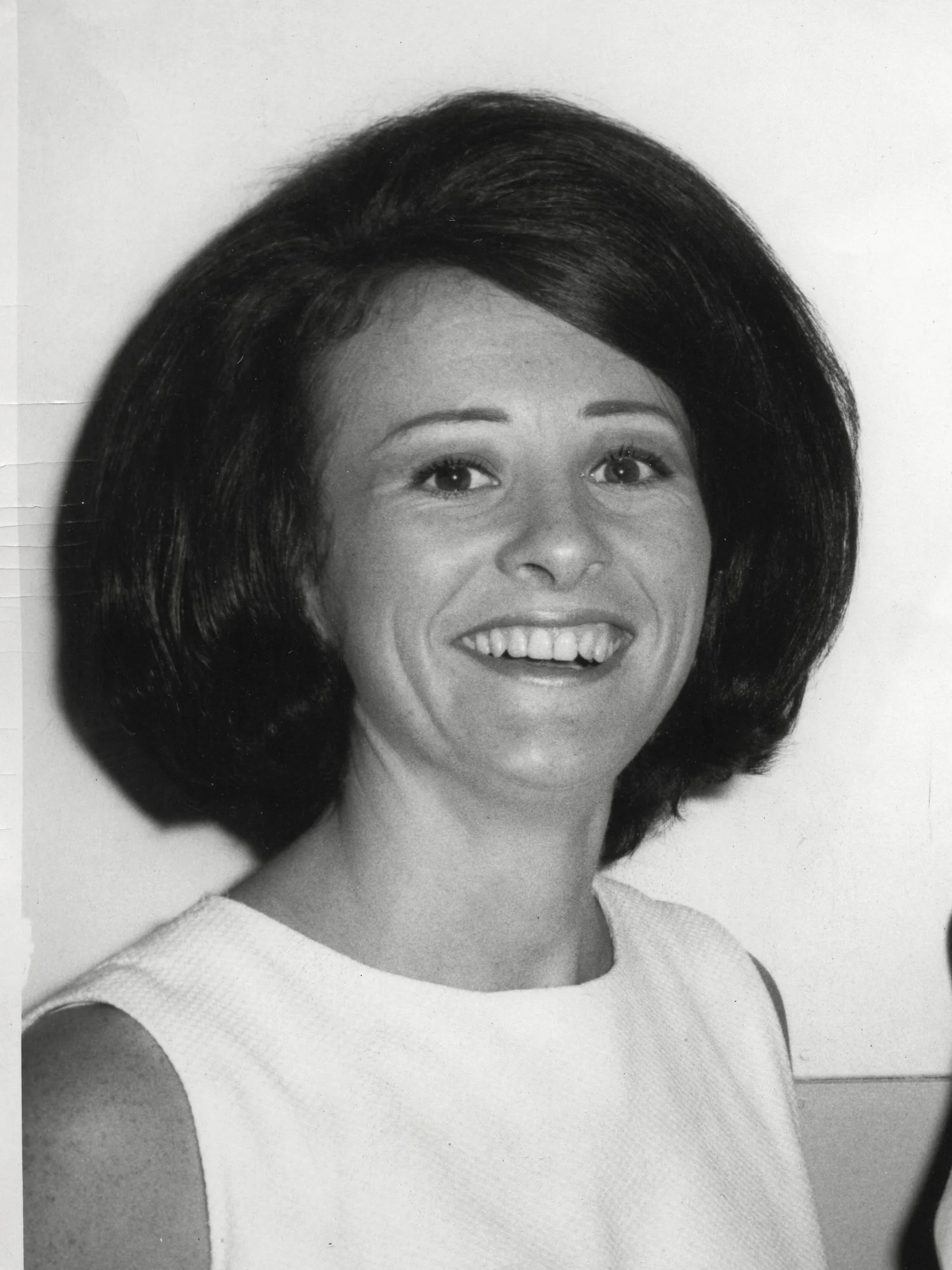 Black and white photograph of a smiling woman with shoulder-length hair, wearing a sleeveless dress, against a plain background.