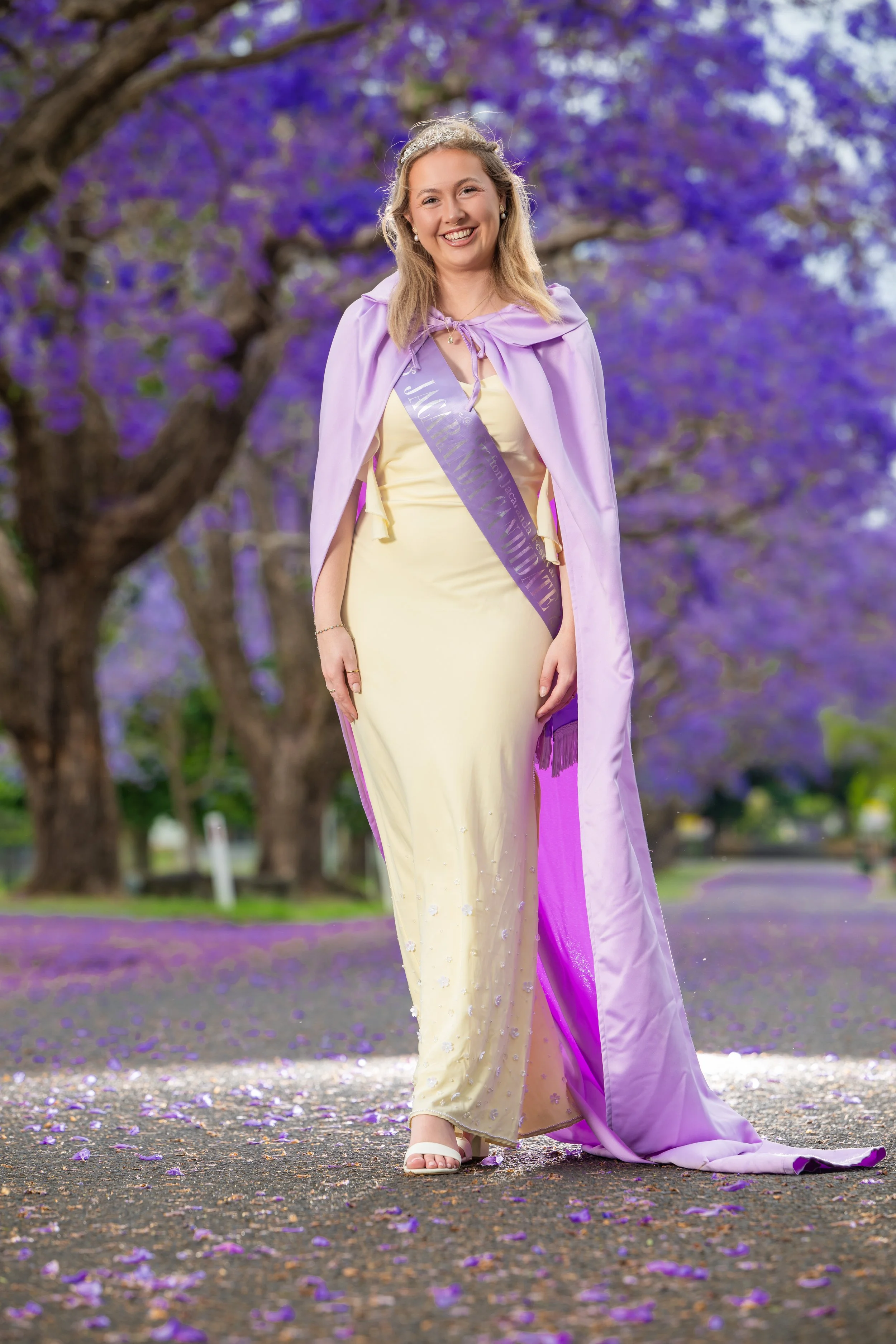 A young woman wearing a sash, tiara, and lavender cape, standing outdoors on a pathway covered with purple flowers. She is smiling and wearing a light yellow dress and white sandals, with purple flowering trees in the background.