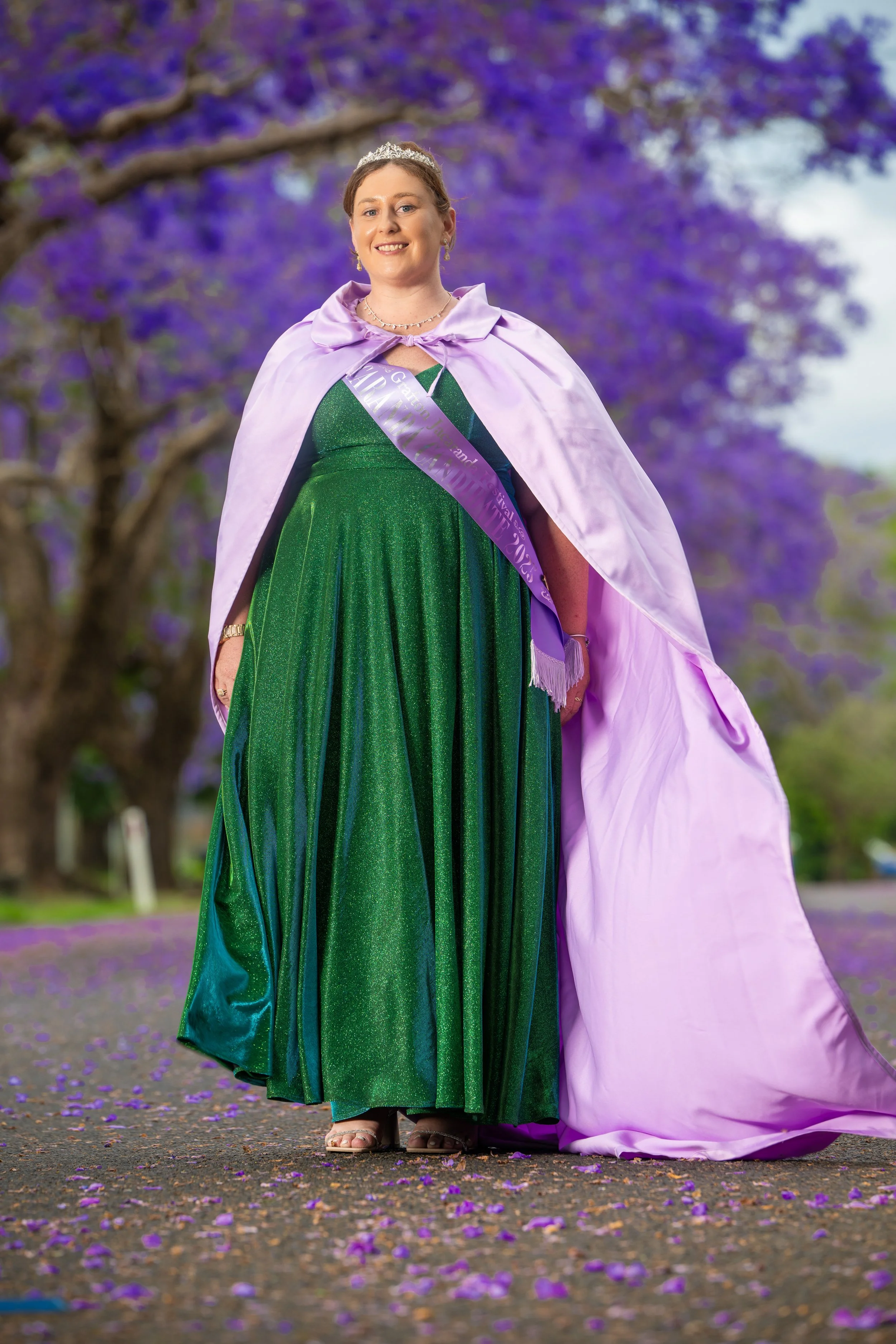 Woman in a green gown and purple cape standing on a pathway with purple flowering trees in the background.