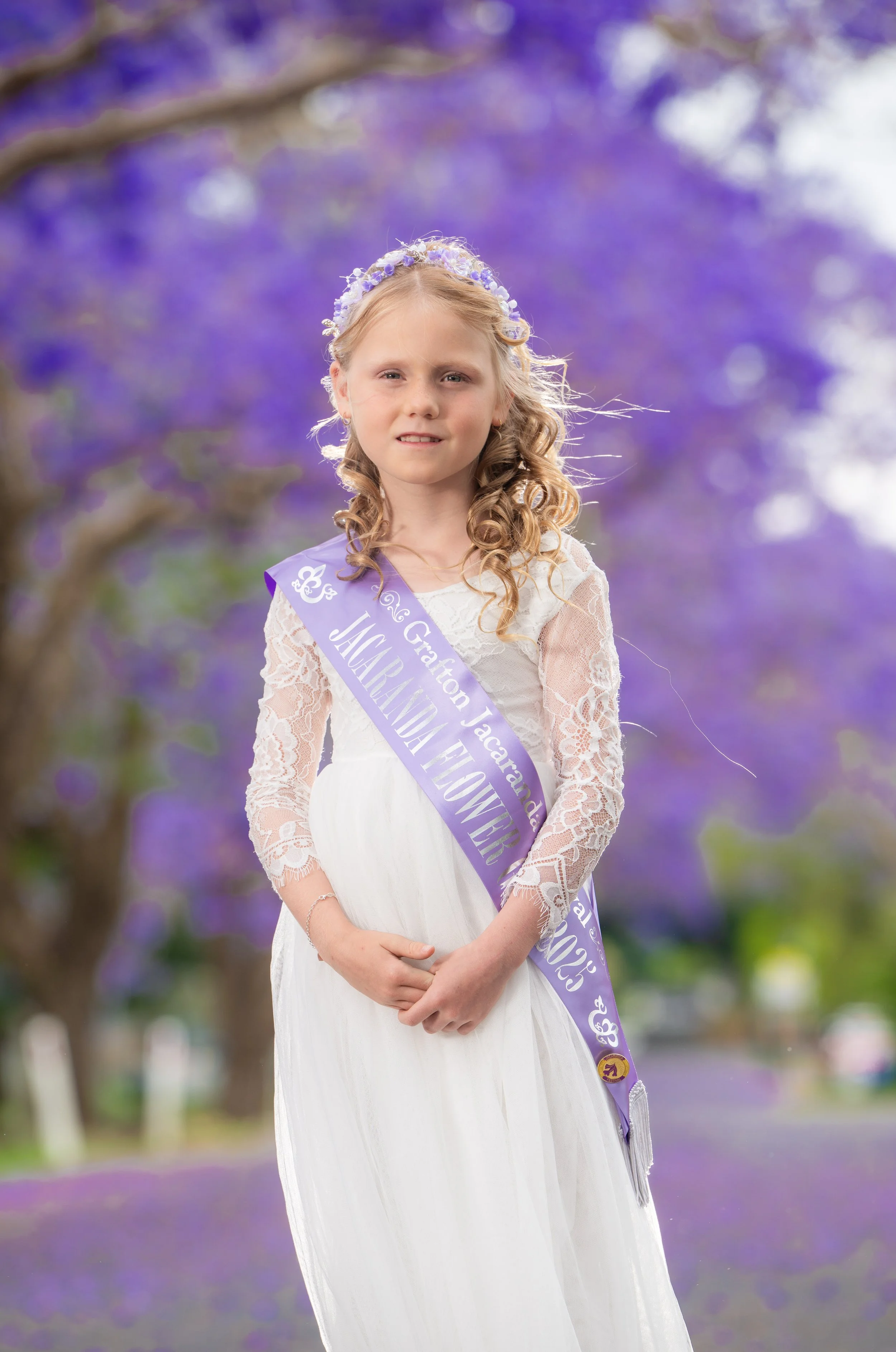 Young girl wearing a white dress and a purple sash that reads 'Prom Queen 2023' with a floral headband, standing outdoors with blooming purple trees in the background.