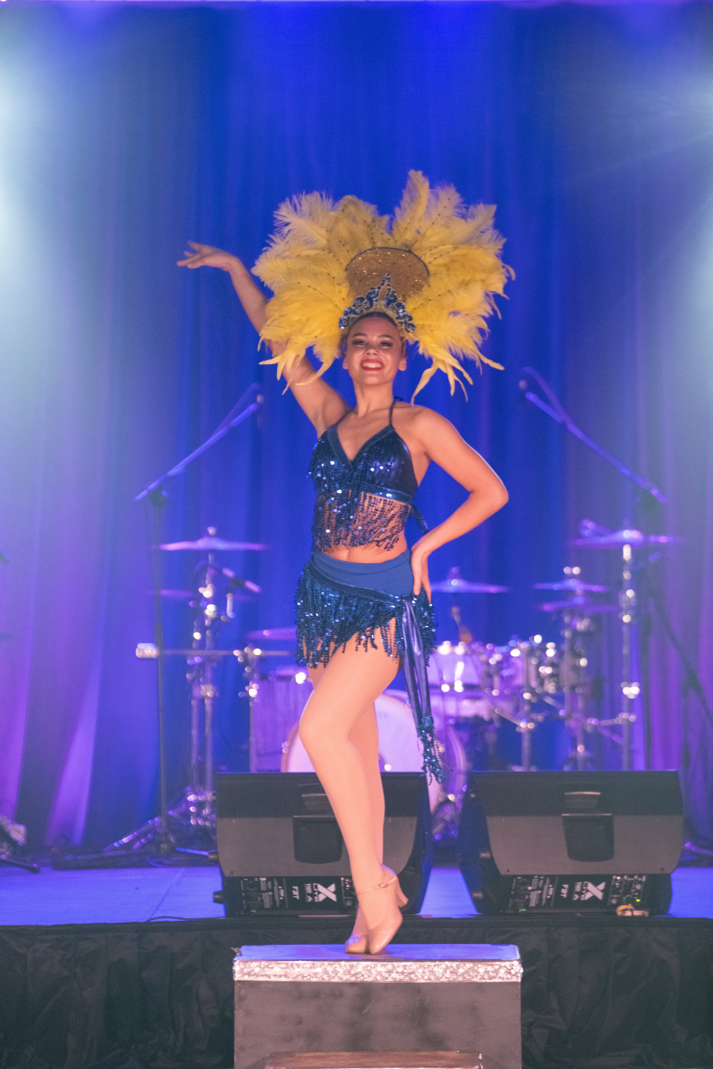 A woman in a black and blue sequined costume with a large yellow feathered headdress performs on stage. She is standing in a pose with one arm raised and smiling brightly. The stage has purple lighting and musical instruments in the background.
