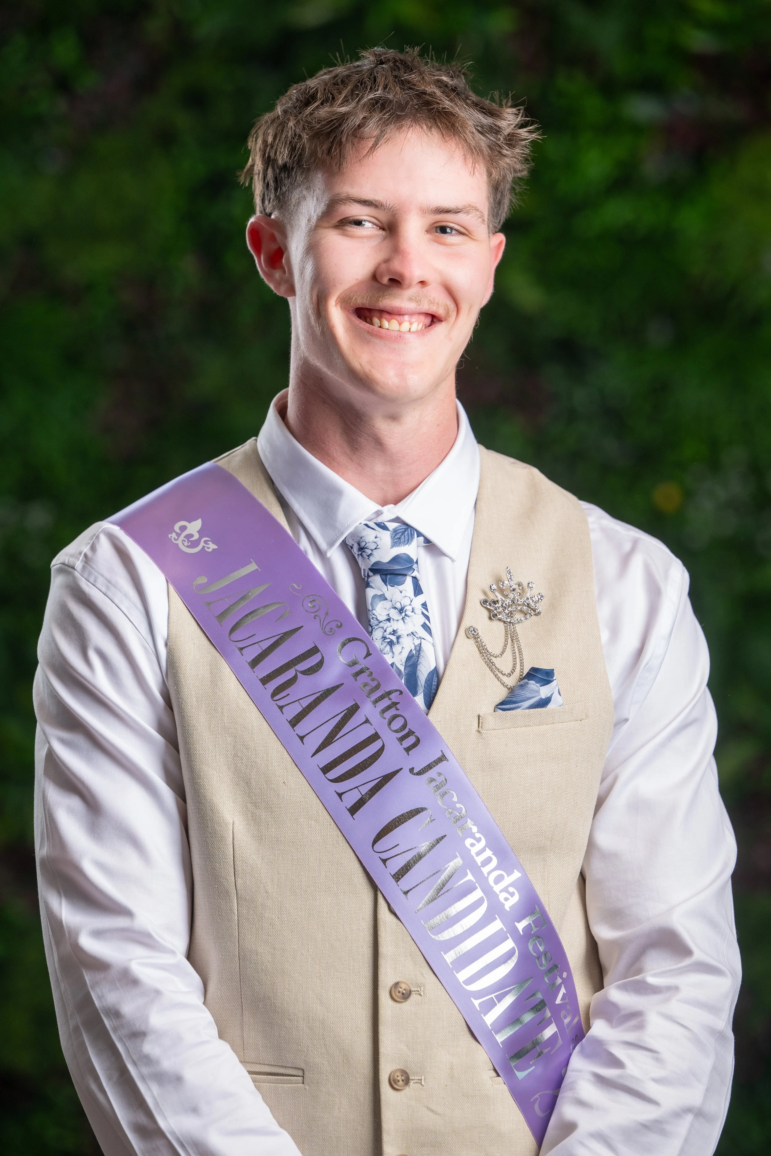 Young man in formal attire smiling, wearing a purple sash with text and decorative pins, standing outdoors with green foliage in the background.