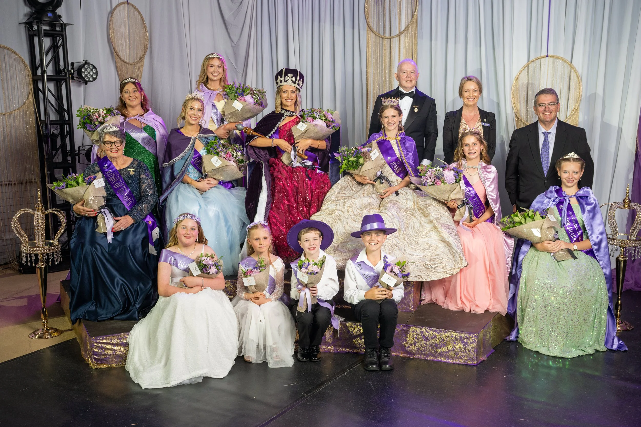 A group of women, girls, and two men in formal and royal costumes posing on a stage with a white backdrop and decorative round gold accents. The women and girls are wearing tiaras, sashes, and holding bouquets, suggesting a pageant or celebration event.