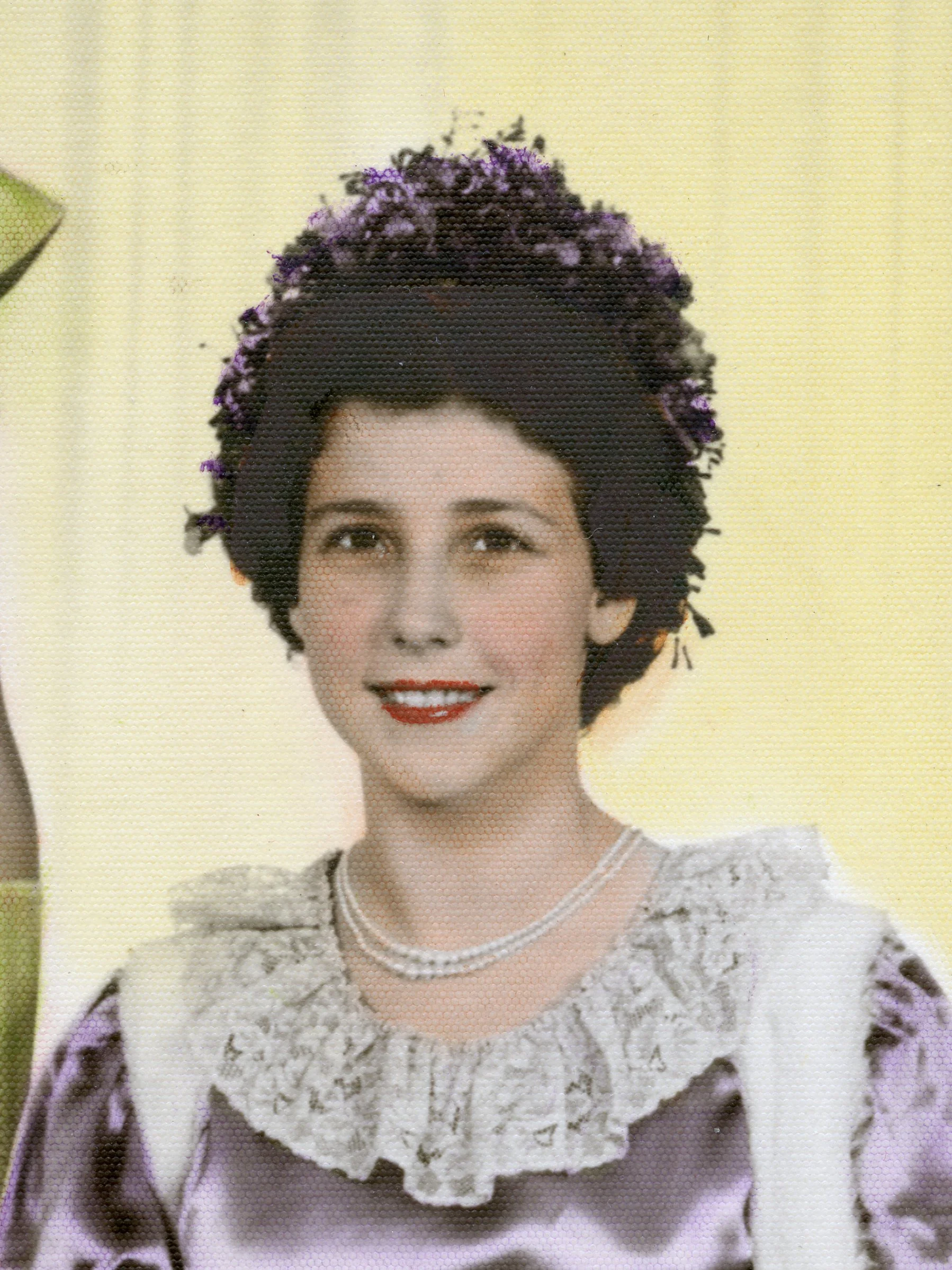 Portrait of a woman with dark, curly hair adorned with purple flowers, smiling, wearing a lace top with a necklace, against a pale yellow background.