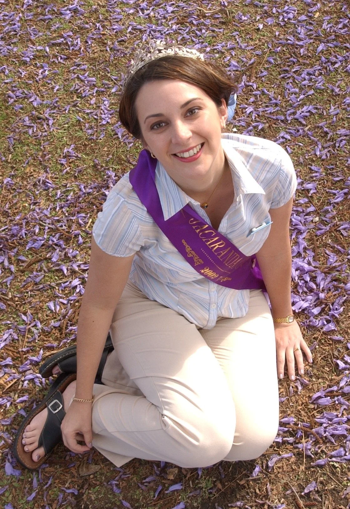 A woman sitting on the ground covered with purple flowers, wearing a crown and a purple sash that reads "Vocaladora 2000".