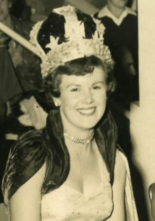 A black-and-white photo of a smiling woman wearing a crown, a lace dress, a pearl necklace, and a cape. She is at a celebration or pageant event, with other people in the background.