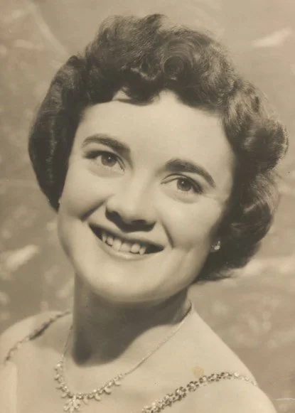 Black and white portrait of a smiling woman with short, curly hair wearing a pearl necklace and earrings.