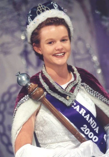 Young woman dressed as a queen with a crown and sash, holding a scepter in a coronation setting.