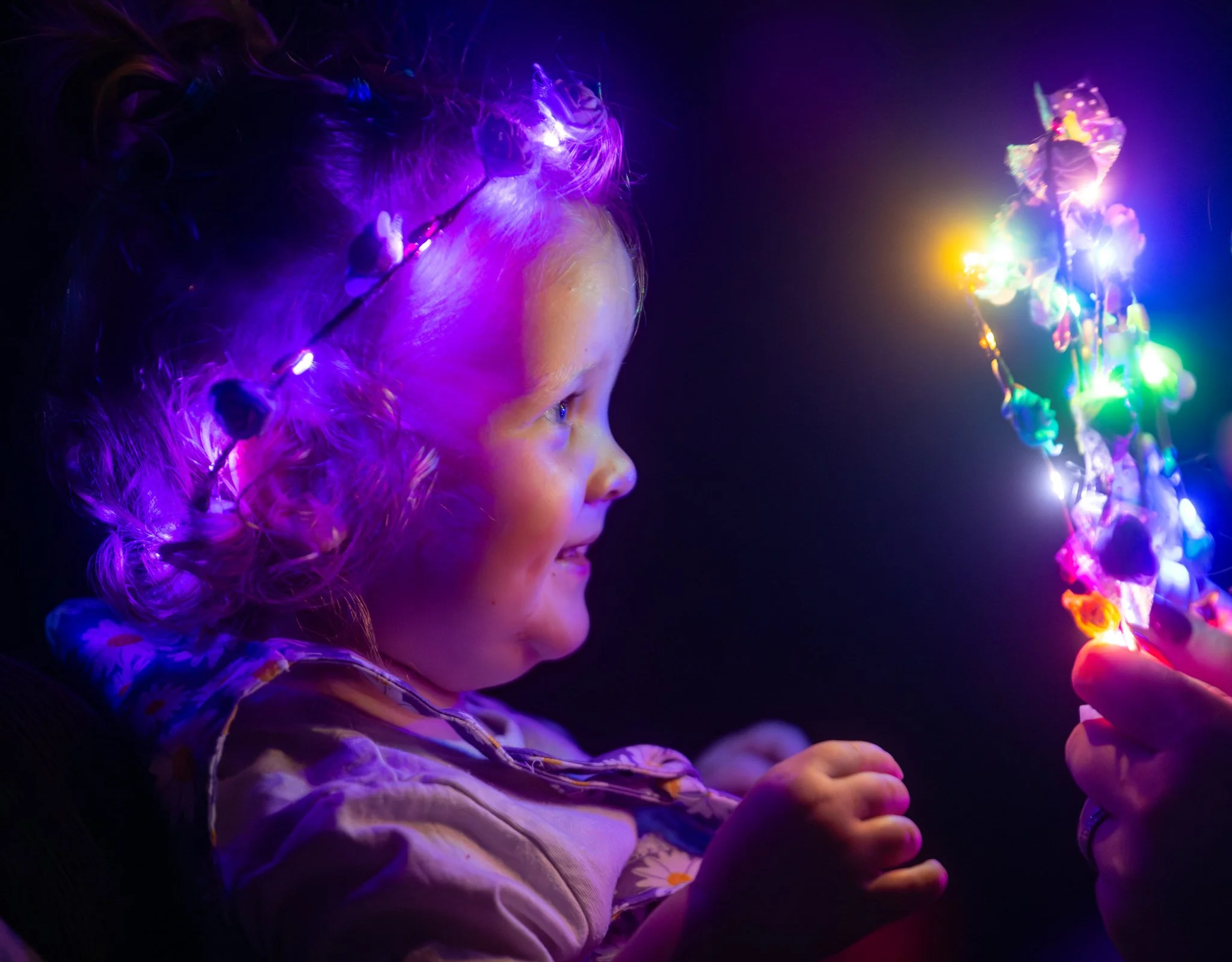 A young girl with curly hair is illuminated by colorful LED lights, looking at a glowing object.
