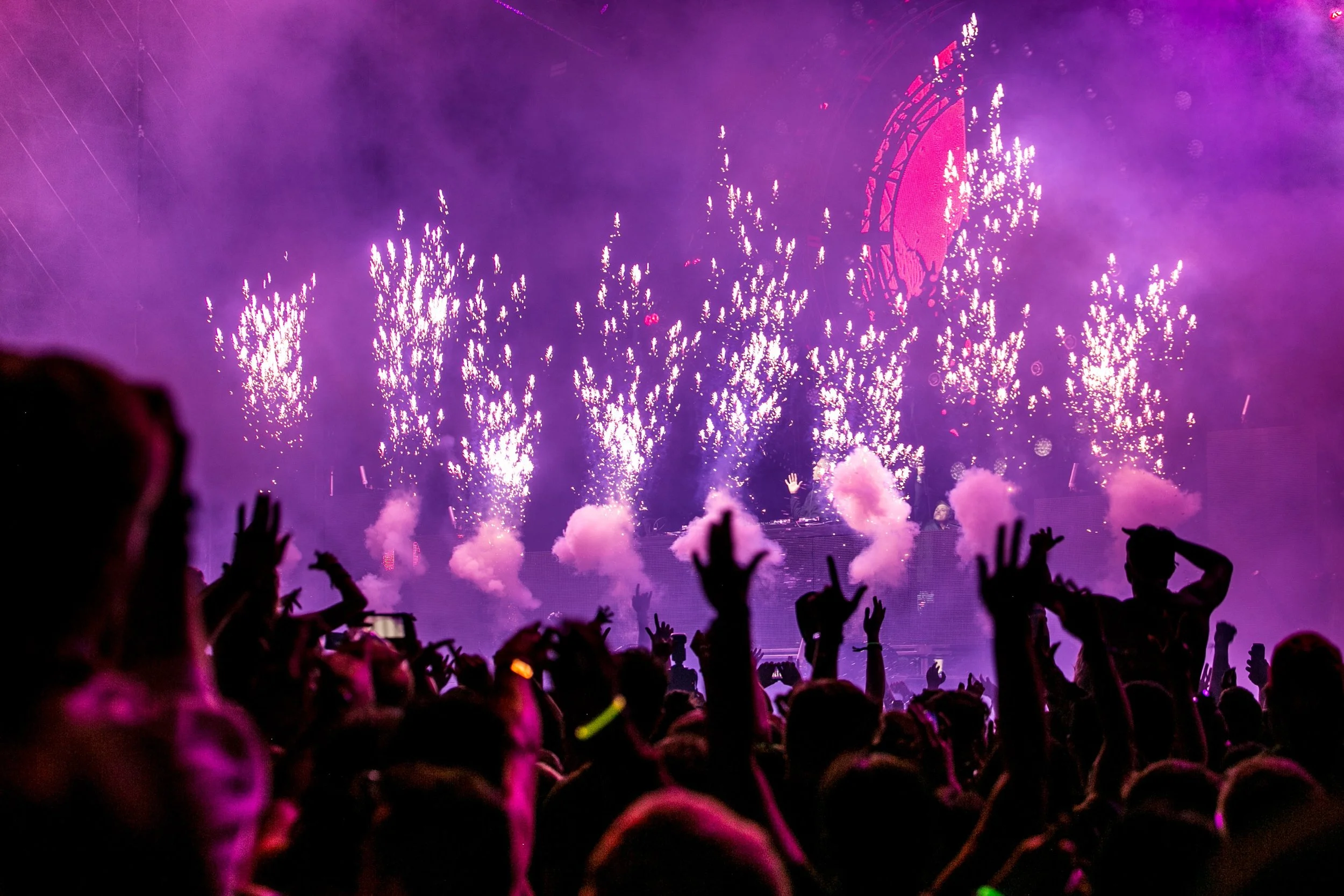 A crowd watching a colorful fireworks display at night with purple and pink lights, and silhouettes of people raising their hands.
