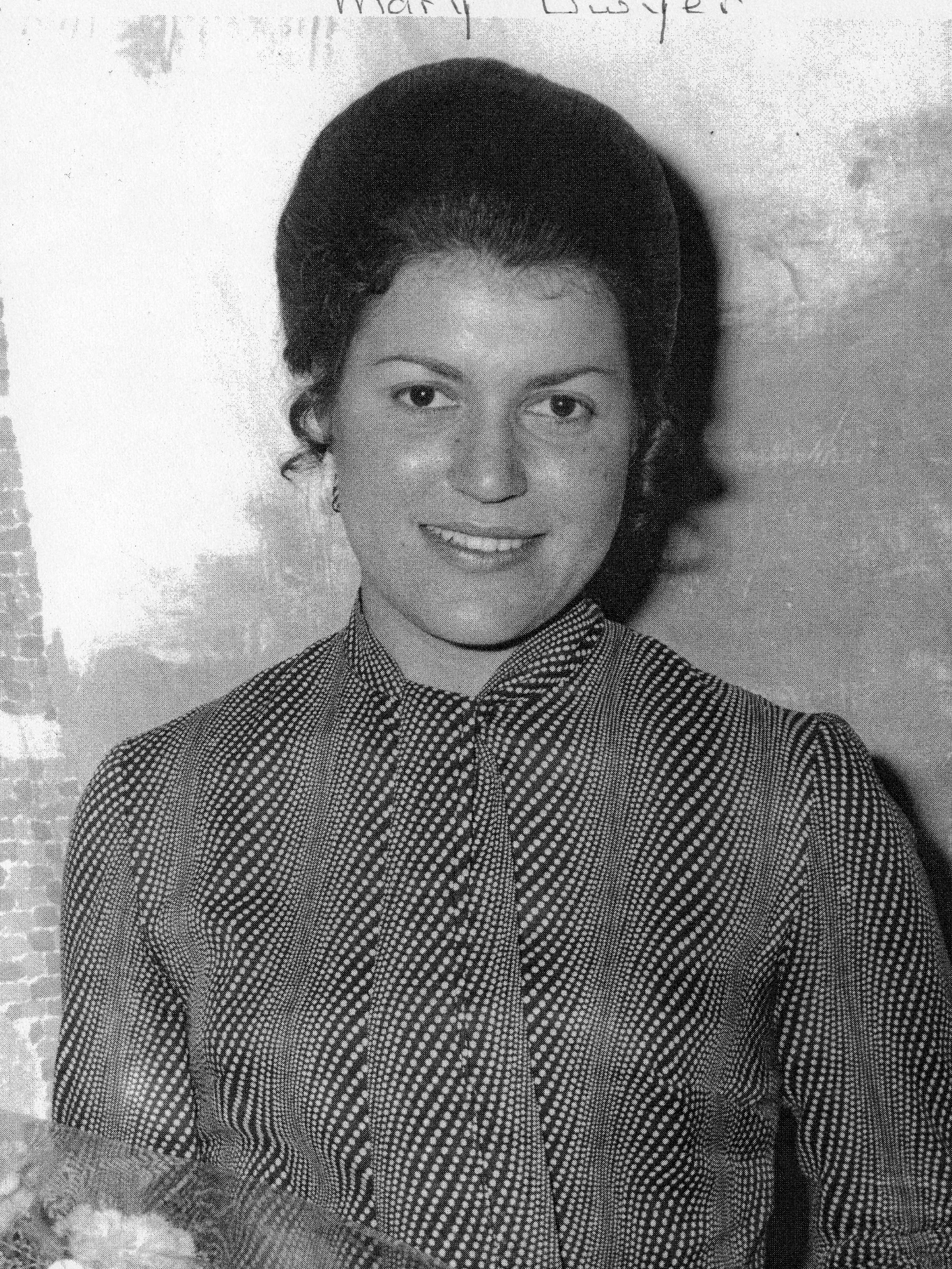 Black and white photo of a woman with dark, styled hair, smiling, wearing a checkered top, standing outdoors near foliage.