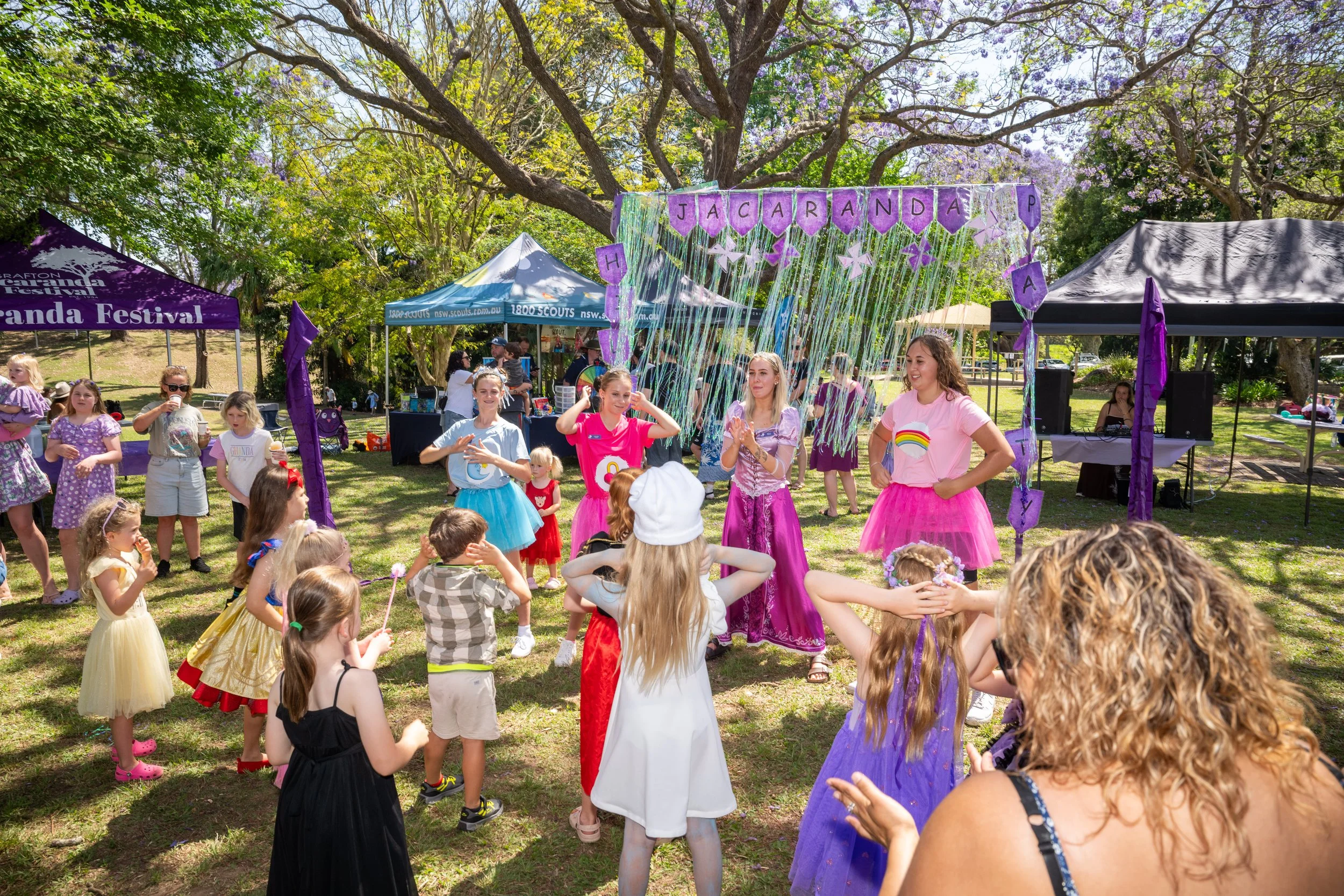 Children and adults participating in a festival in a park with trees and purple decorations, including a banner that says "JACARANDA" and various tents.