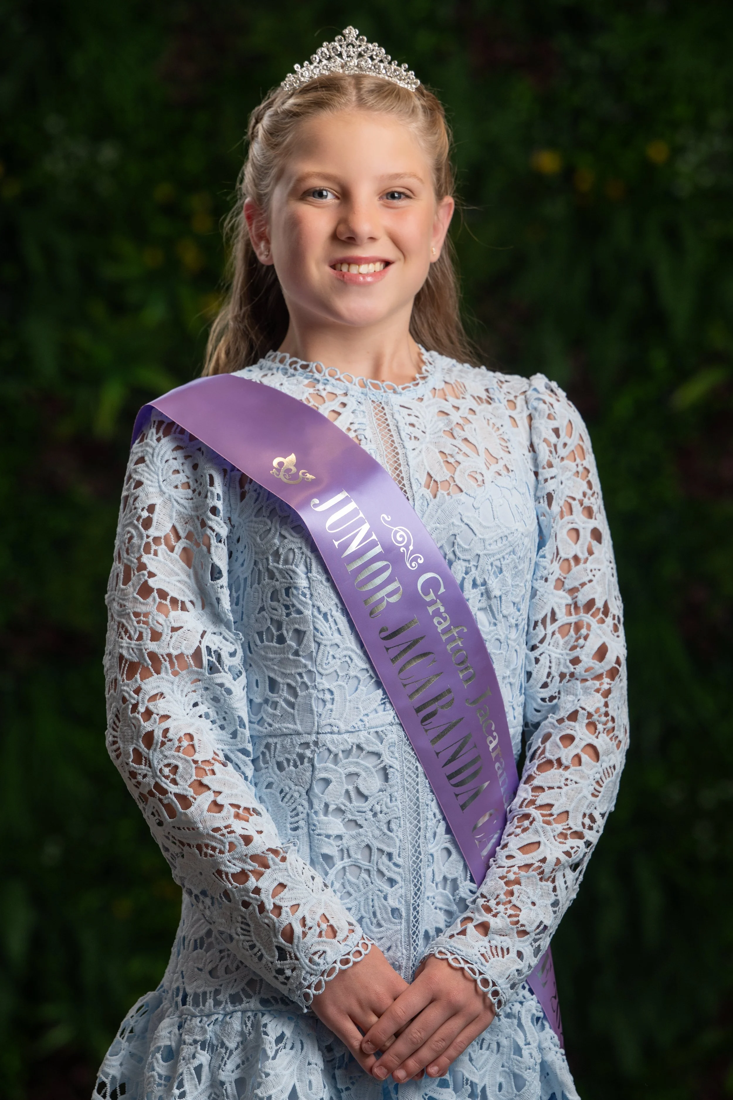 Young girl wearing a crown and sash, dressed in a lace white dress, smiling with hands folded in front of her, outdoors with greenery background.