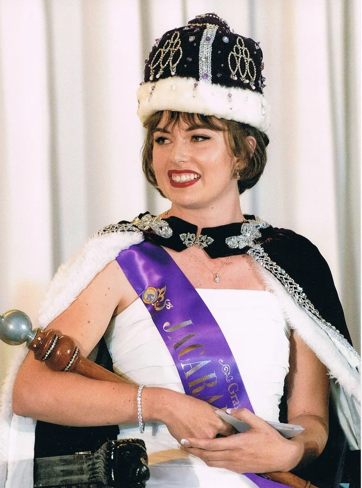 A woman dressed as royalty, wearing a crown and a sash, holding a scepter and smiling.