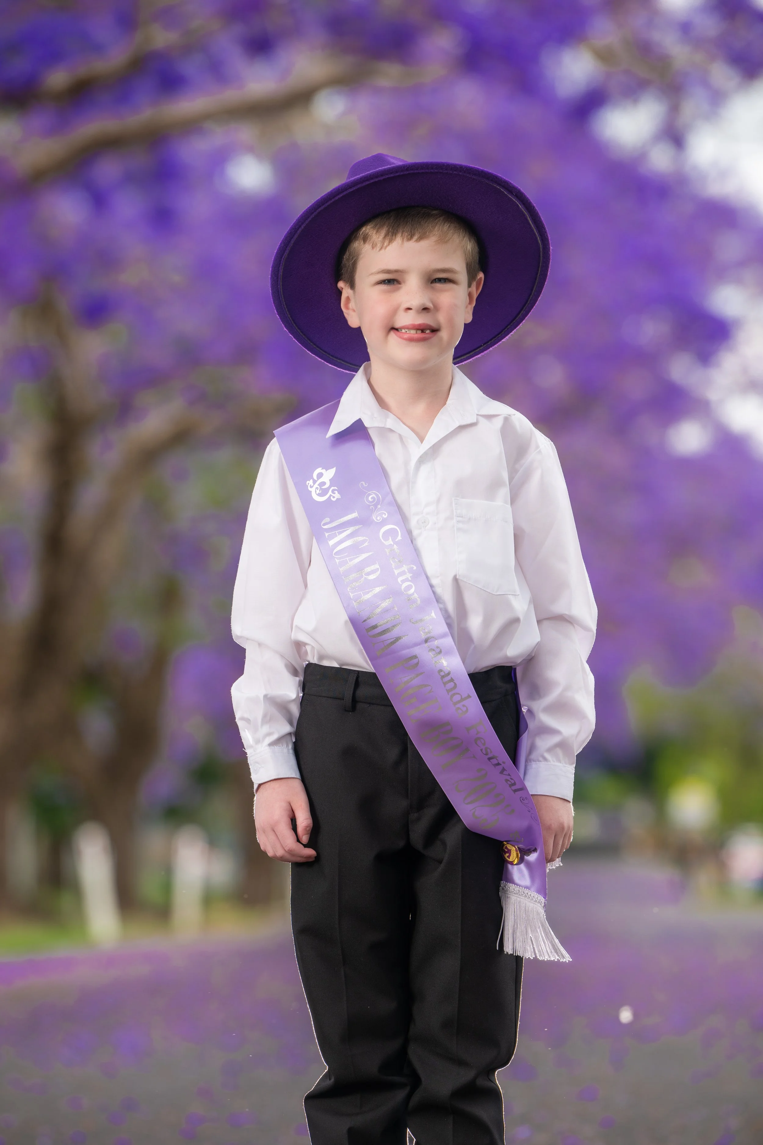 A young boy wearing a purple sash and hat stands in a park with purple flowers in the background. The sash reads 'Jamboree' and other text indicating a festival or celebration.