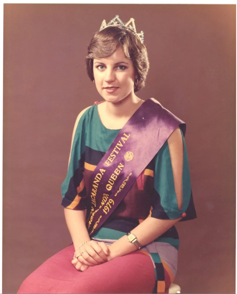 A young woman wearing a crown and a sash that reads 'VIRGINLUANDA FESTIVAL 1979 QUEEN' in gold letters, sitting with her hands folded on her lap against a plain brown background.