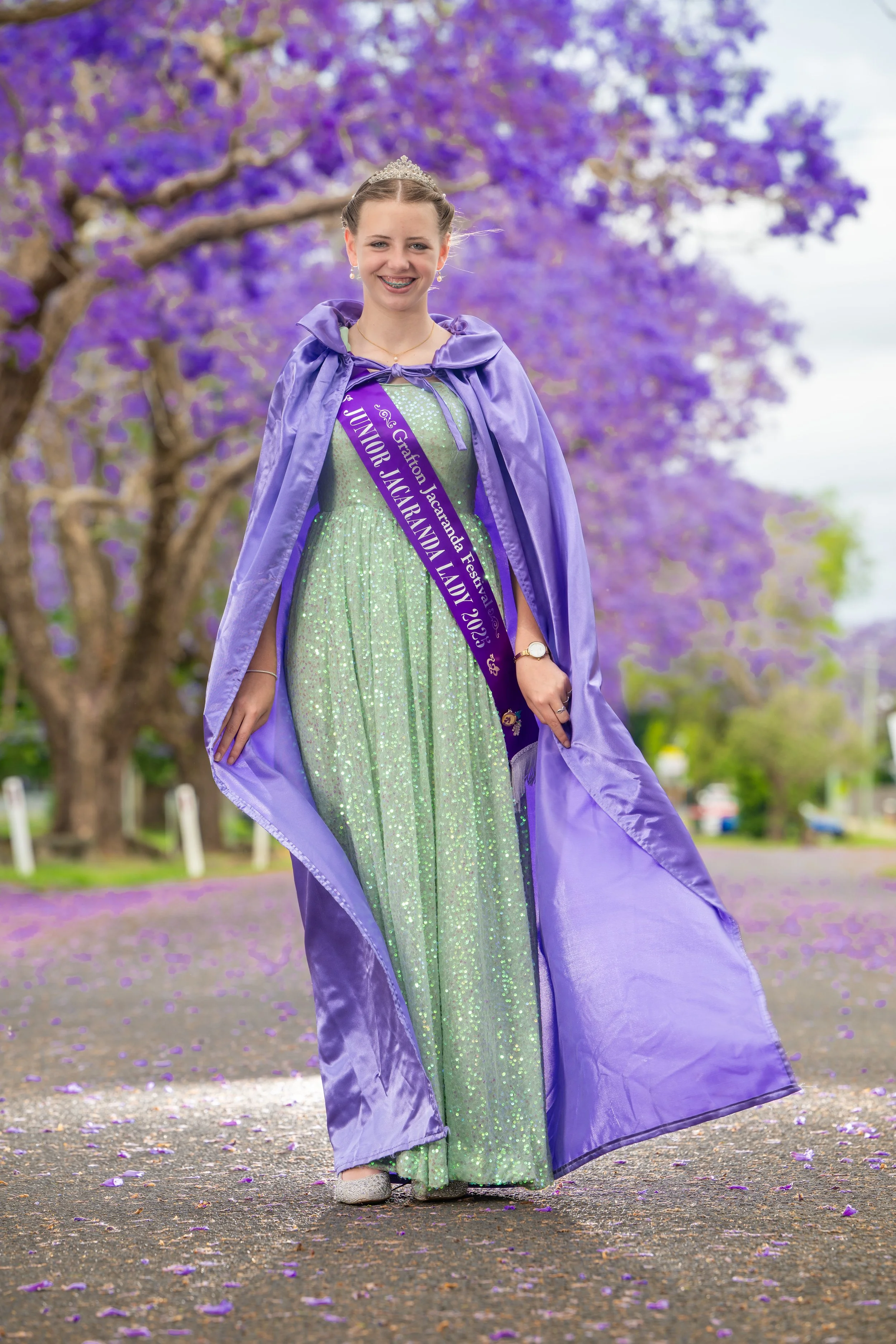 Young girl in a sparkly green gown, purple cape, and tiara, smiling outdoors with purple flowering trees in the background, wearing a sash that reads 'Junior Jacqueline Festival 2023'.