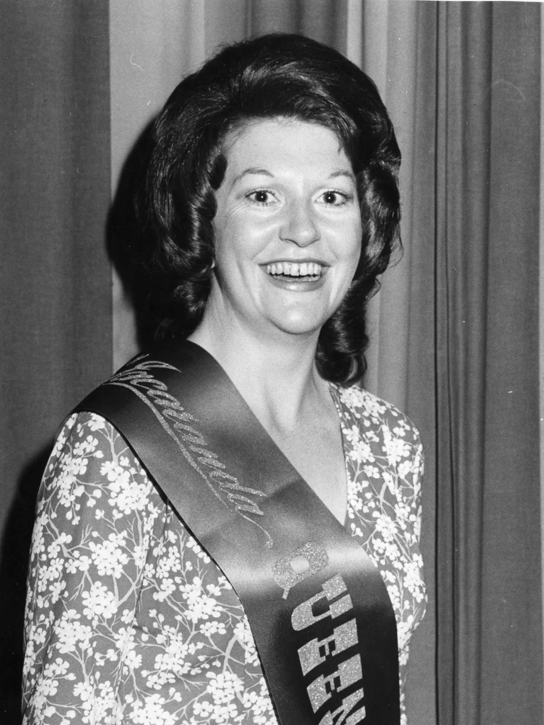 Black and white photograph of a woman with dark, voluminous hair and a wide smile, wearing a floral blouse and a sash that reads 'Miss America.'