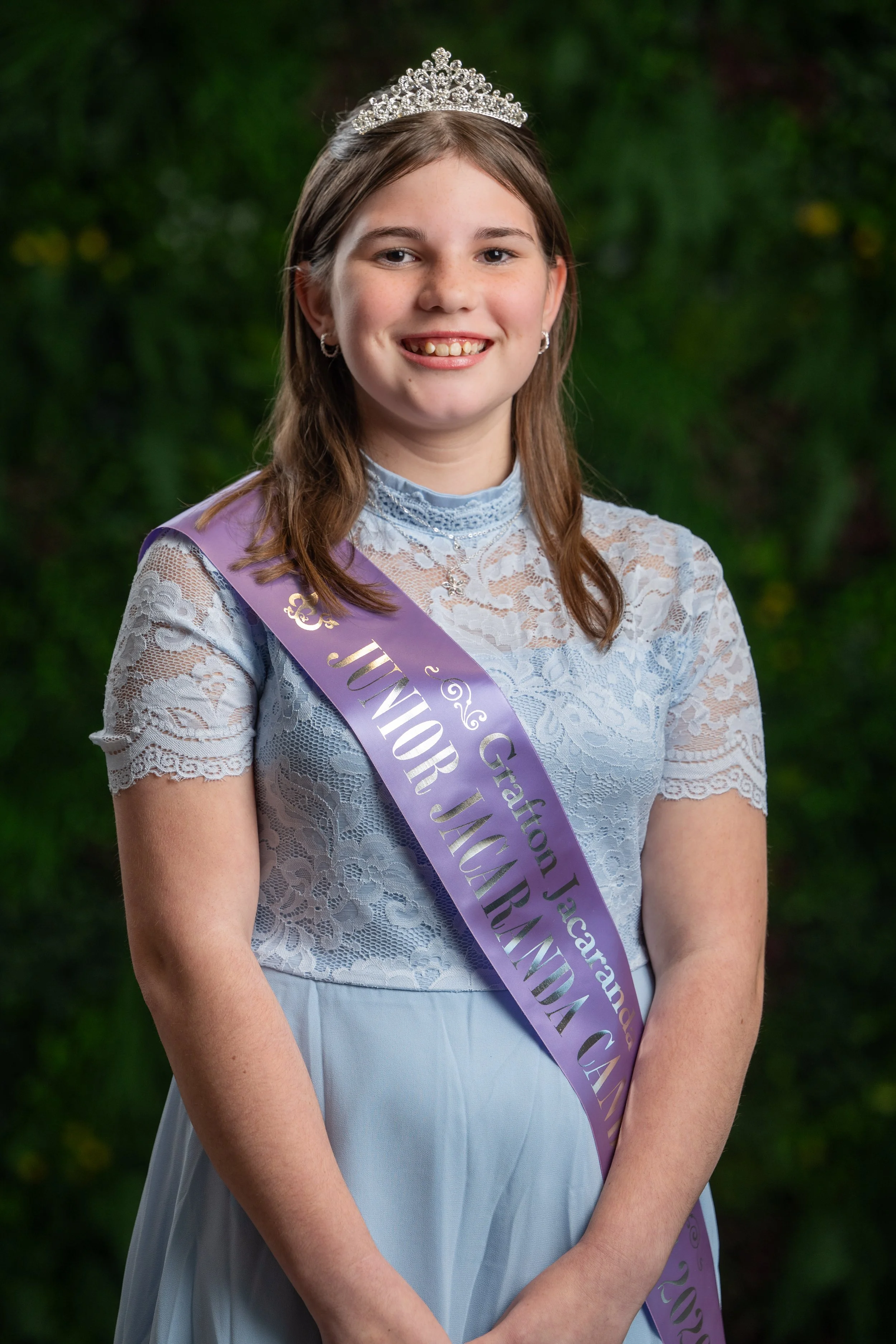 Young girl with brown hair wearing a tiara, earrings, and a light blue lace dress, standing outdoors with greenery in the background. She is smiling and has a purple sash across her chest that reads 'Junior J'.