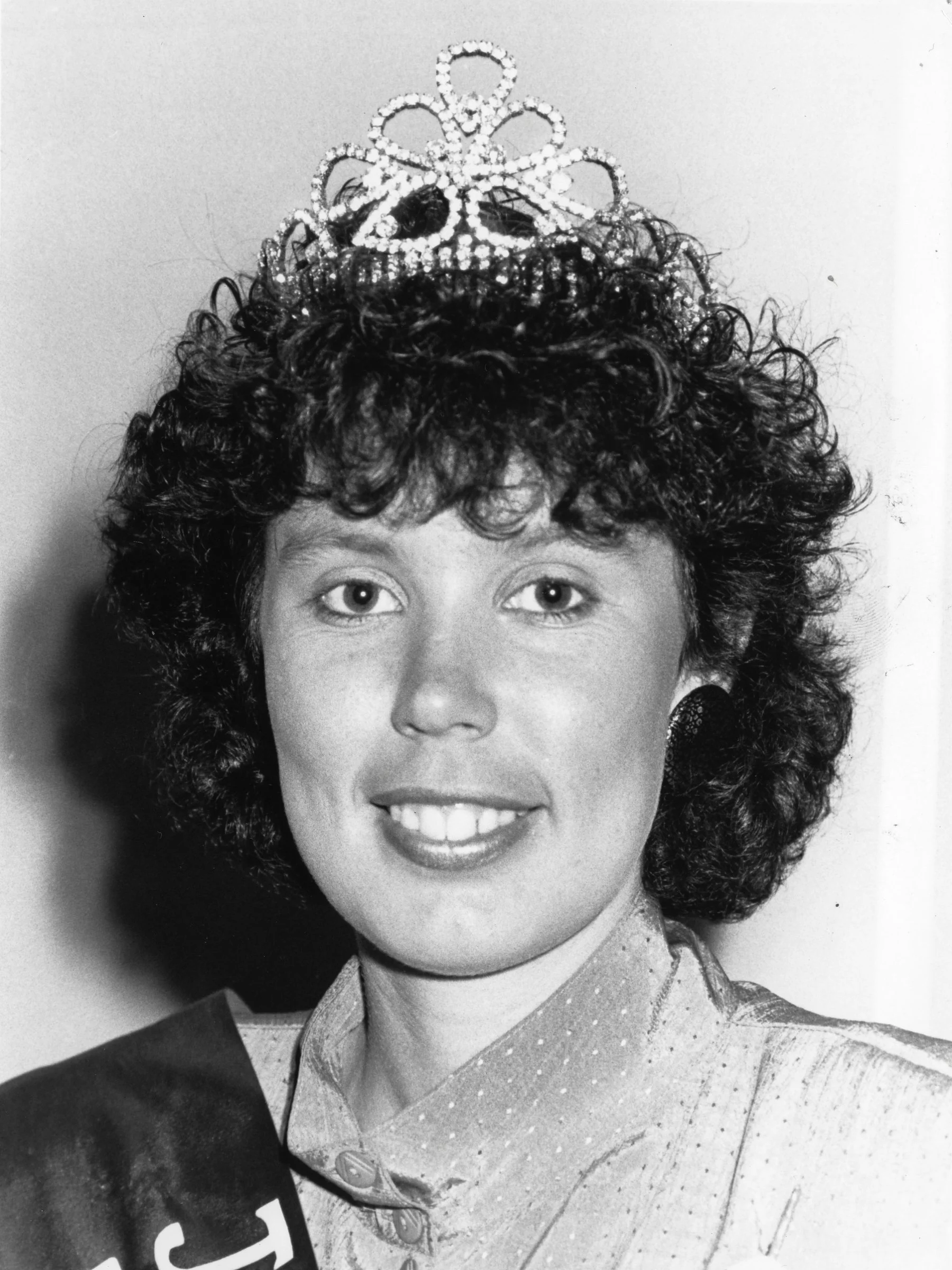 Black and white photograph of a young woman with curly hair, wearing a dress with a collar, and a small tiara on her head.